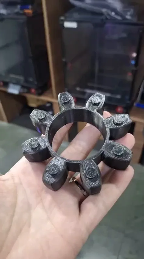 Peça preta impressa em TPU sobre a mão, com impressoras Voron ao fundo